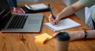 Person macht sich mit Bleistift Notizen neben Laptop und Taschenrechner am Schreibtisch